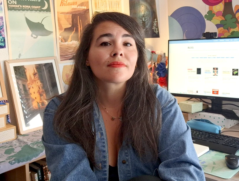 Femme brune avec une veste en jean bleu regarde la caméra. Derrière elle, un écran d'ordinateur allumé sur un blog, un bureau en beau et un mur recouvert de prints colorés.